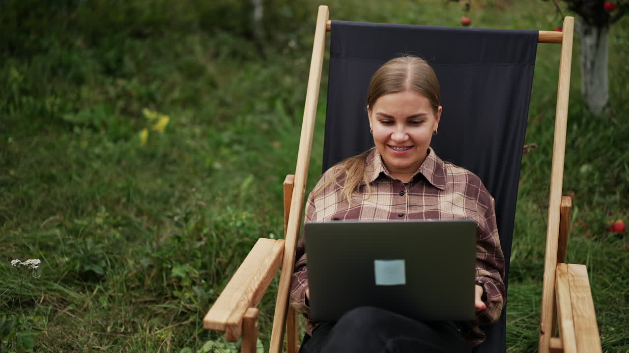 Positive smiling young woman sits in garden chair working on laptop. Lady distracts for a moment, closes her eyes to have a little break from remote work.