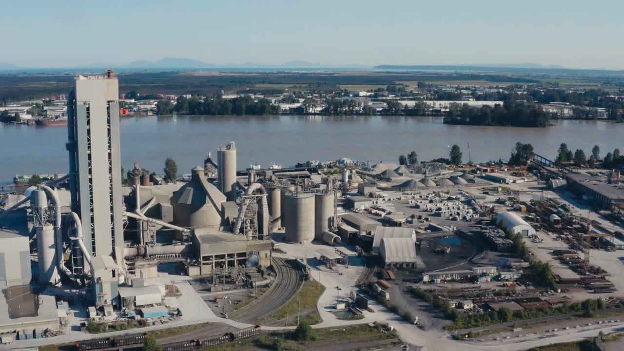 Aerial fly over cement refinery factory that pipes raw materials of oil gas metal through chimney exhausts and exports off the port with freightors docked loaded and reasy to transport the raw product