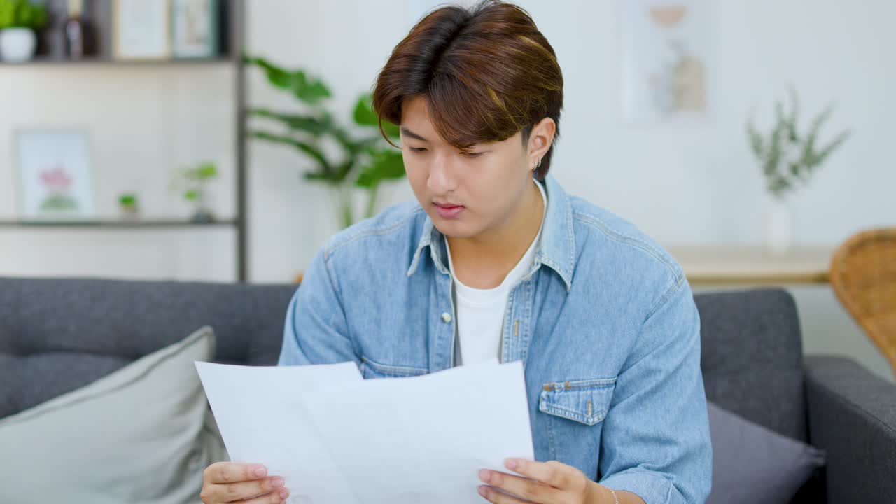 デニムシャツを着た若いアジア人男性が、室内で書類を丁寧に確認しています。