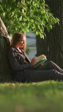 mujer sentada al aire libre con las piernas extendidas, apoyada en un árbol en un campo cubierto de hierba, hojeando las páginas de un libro, las hojas de los árboles balanceándose suavemente en la brisa, y la vegetación en el fondo