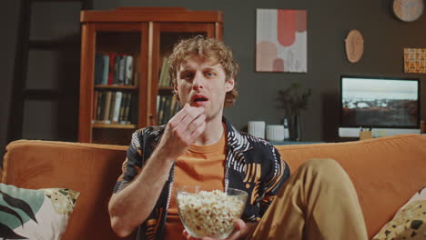 portrait of man sitting on sofa, eating popcorn and smiling as watching tv