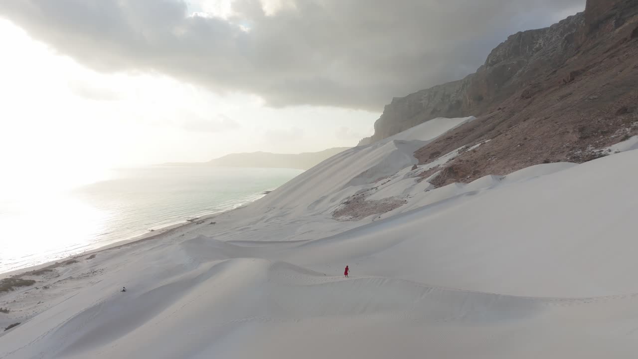 воздушный вид одинокой фигуры в красном, идущей по белым песчаным дюнам рядом с пляжем арчера во время восхода солнца в сокотре