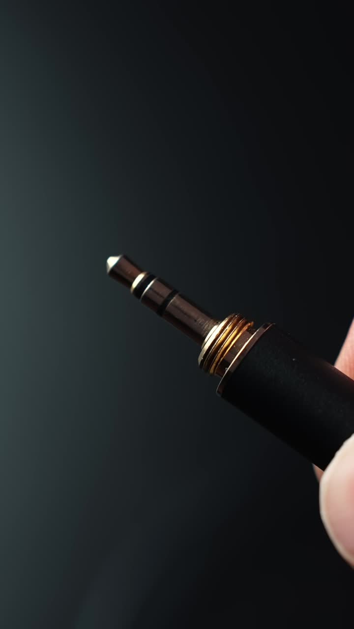 Macro a fuoco morbido di un jack aux da 3,5 mm tenuto dalle dita su uno sfondo scuro con spazio per il testo, punti luce caldi e un'atmosfera tecnologica minimale e suggestiva.
