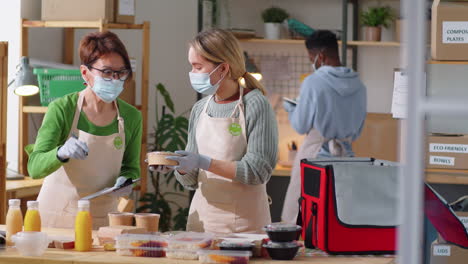 mujeres con máscaras empaquetando contenedores de alimentos en bolsas de entrega