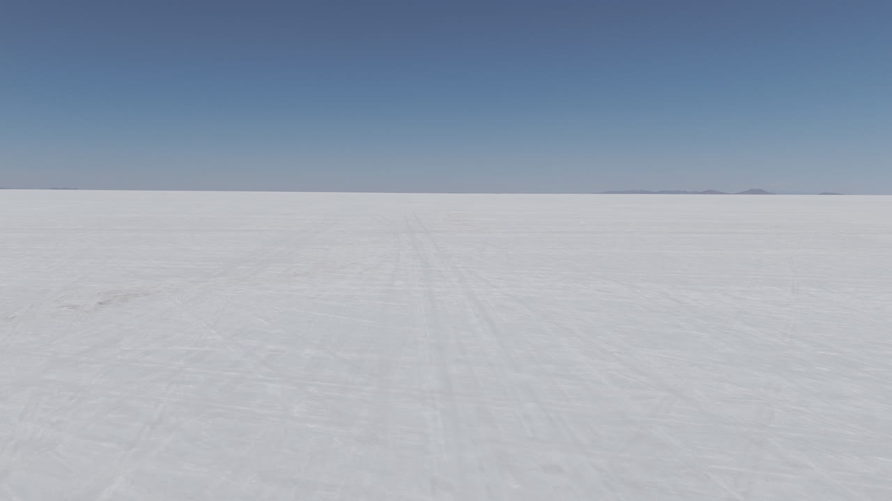 Drone shot flying above the dry salt flats in Bolivia Uyuni on the cloudy day with some small hills in the background and car tracks in the salt LOG