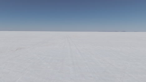 drone shot flying above the dry salt flats in bolivia uyuni on the cloudy day with some small hills in the background and car tracks in the salt log
