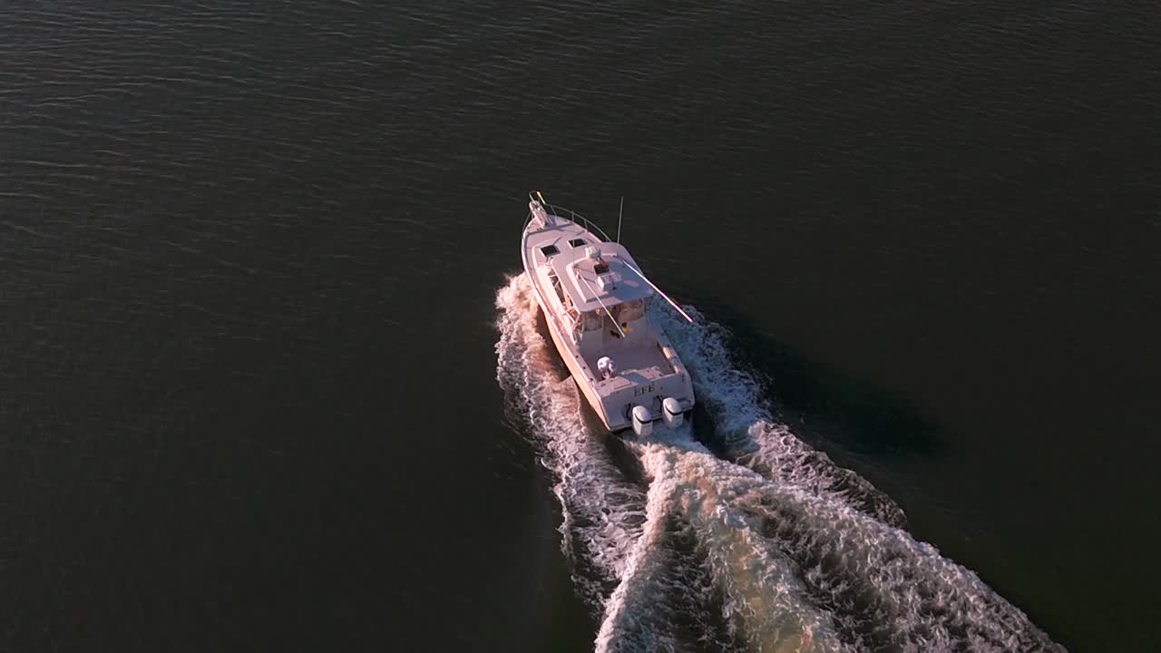 An aerial view, behind a white fishing boat in the bay off the southern shores of Long Island, NY on a sunny day. The camera dolly in behind the vessel, as two people in the boat enjoy the day