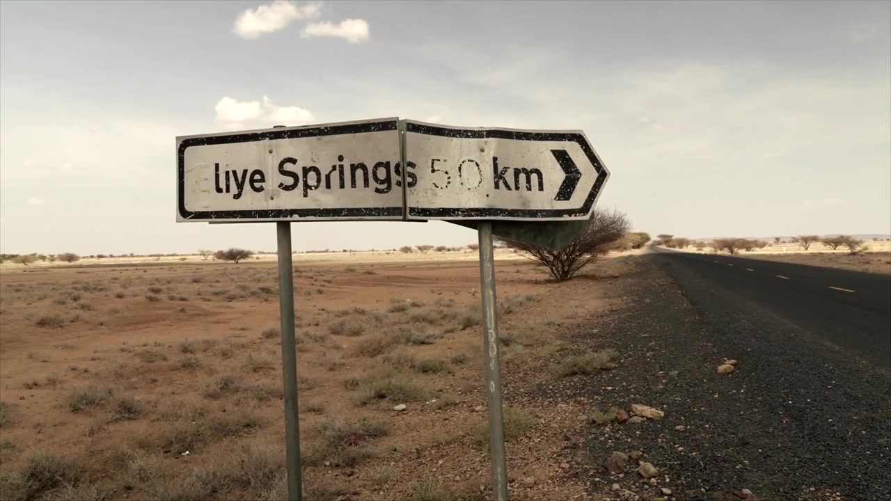 Segnale stradale usurato che indica Eliye Springs a 50 km, in piedi accanto a un'autostrada asfaltata vuota nel vasto e arido paesaggio della contea di Turkana, Kenya, sotto un cielo nuvoloso.