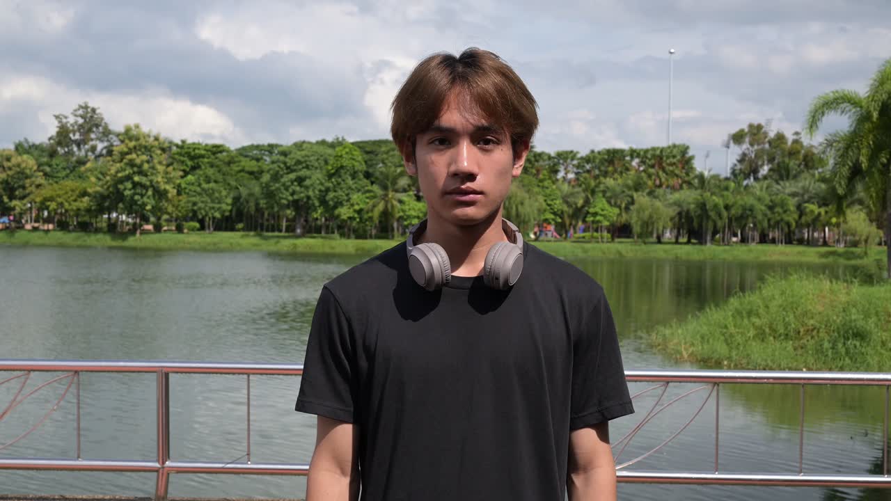 Young Asian handsome man wearing black t-shirt and headphones in park during summer and looking at camera while smiling