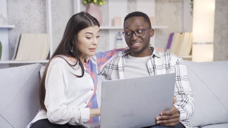 una pareja casada amorosa multiétnica mirando la computadora portátil en la sala de estar, sonriendo, trabajando desde casa, freelance.
