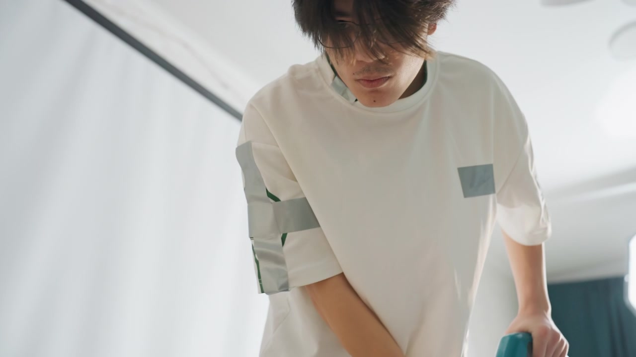 Young man leaning forward indoors preparing home cleaning, soft morning light, curtain backdrop, casual sweater, focused posture, closeup on hands and shoulders, calm preclean routine