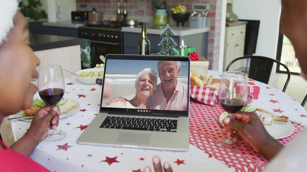 una pareja afroamericana con vino usando una computadora portátil para una videollamada de navidad con una pareja feliz en la pantalla