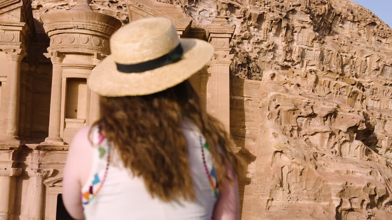 vista trasera de una mujer con sombrero de paja de pie frente al monasterio de ad deir al anochecer en petra, jordania