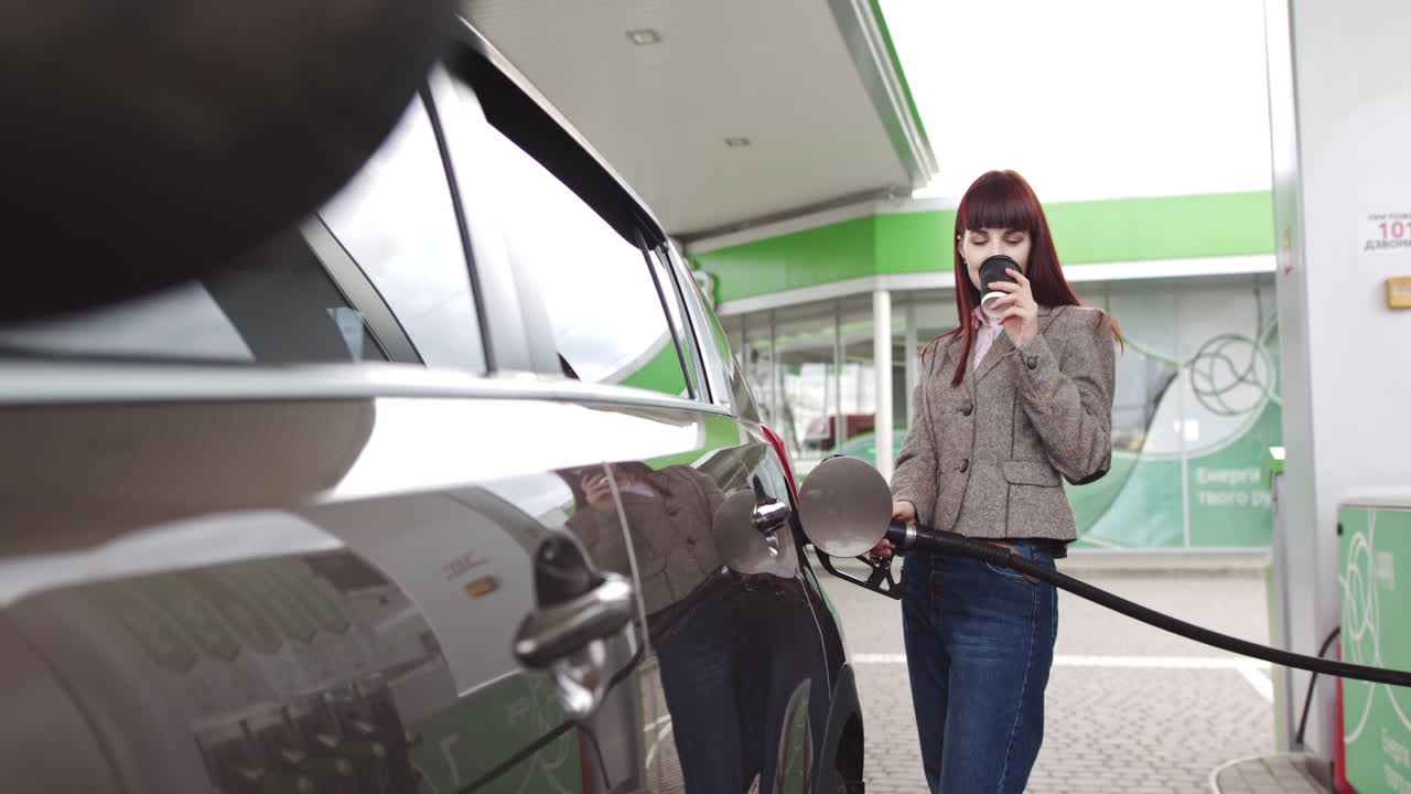Woman filling up her car at a gas station and drinking coffee