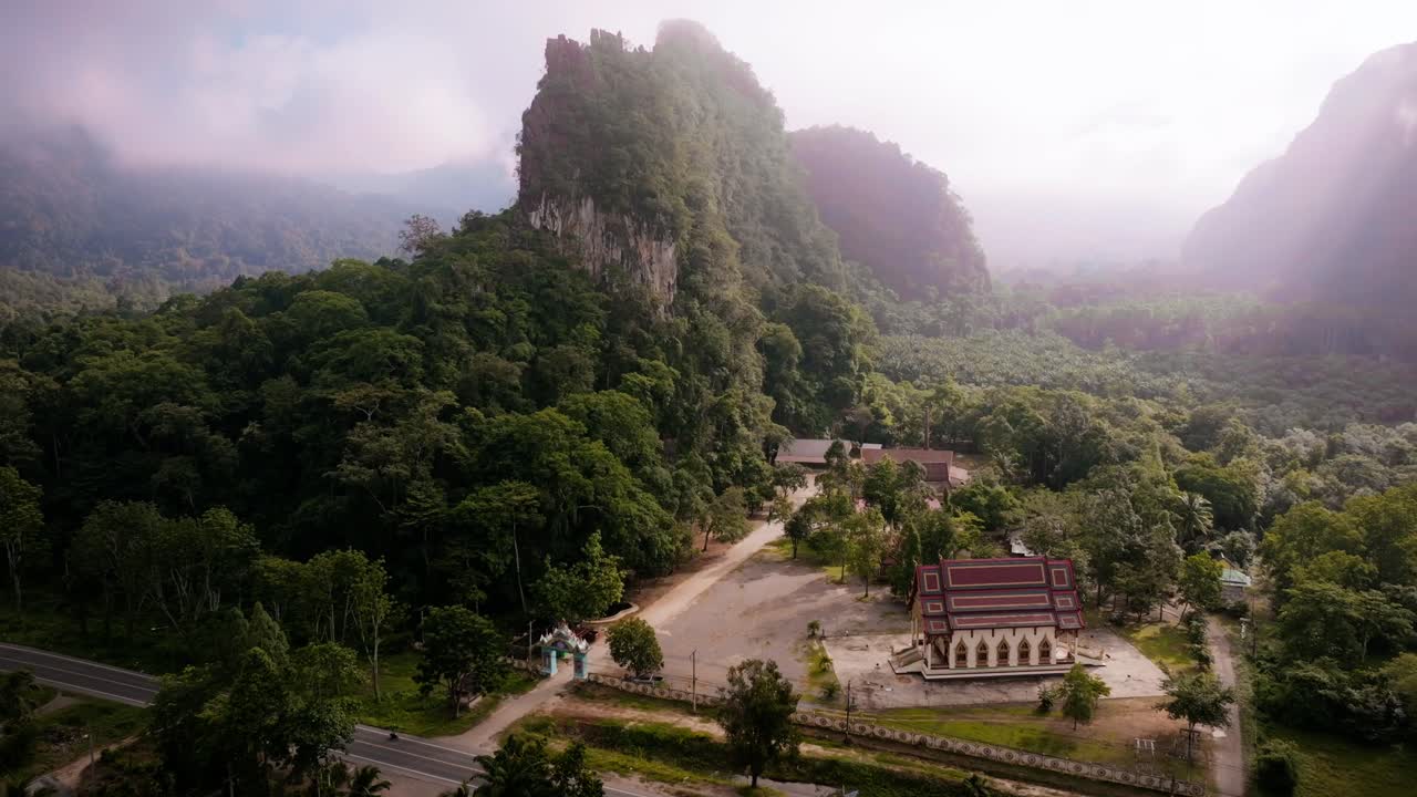 Drone footage of a peaceful Buddhist temple surrounded by jungle and karst mountains in Khao Sok, Thailand. Shot at sunrise with morning mist and lush green scenery