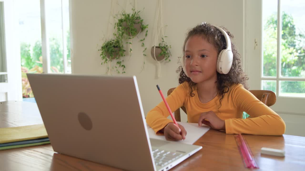 Female child at wooden table, prompted by laptop question, writing with pencil and raising finger