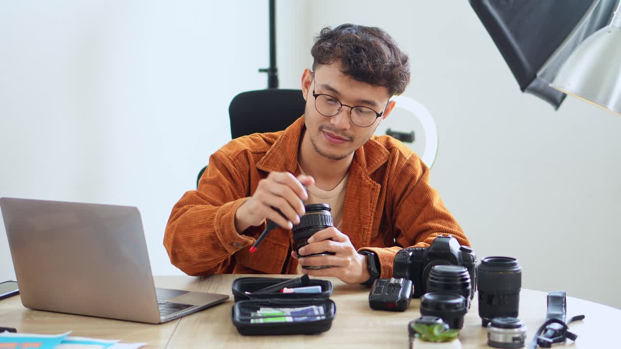 Young Asian Man Wearing Glasses Carefully Cleans DSLR Lens, Preparing His Professional Photography Gear