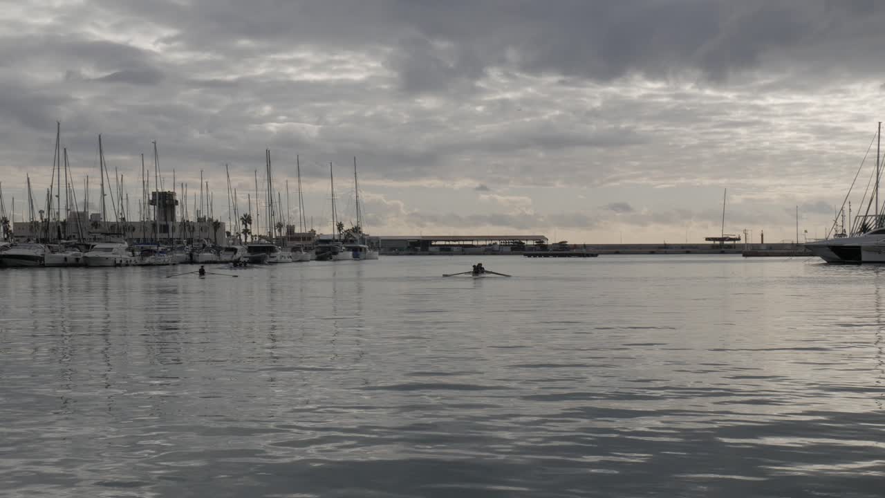 A group of rowers on a calm Alicante harbor, framed by cloudy skies and moored boats