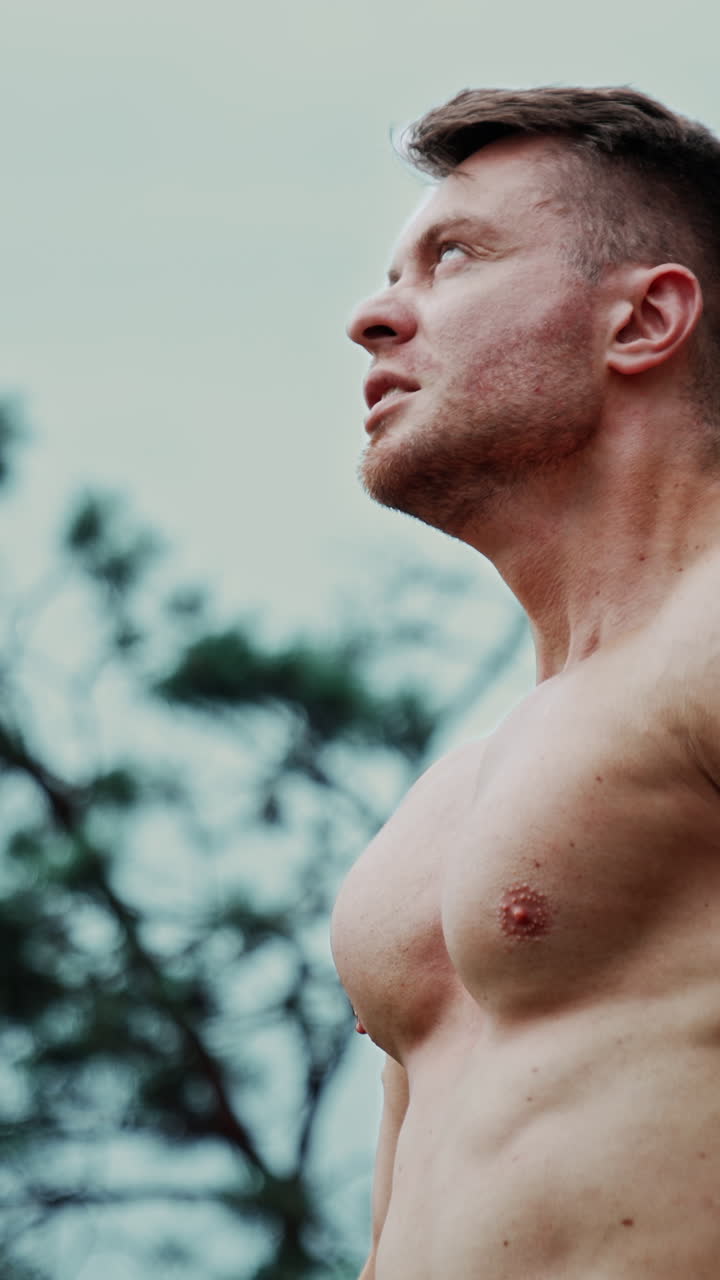 Athletic Caucasian man with naked torso breathing hard after workout. Sportsman exercising outdoors. Low angle view. Vertical video