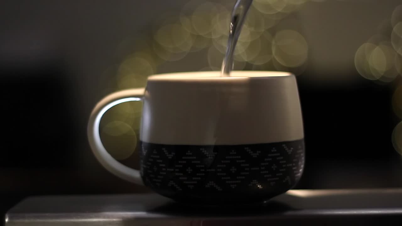 Hot water being poured into beautiful mug with blurred lights illuminating the dark background. Making tea during the cold season. Bokeh videography with shallow depth of field.