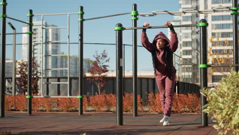 young woman gripping metal bar with both hands while leaning forward under bright sun, hood up, intense gaze on determined face, urban park pull up exercise showing strength and resilience