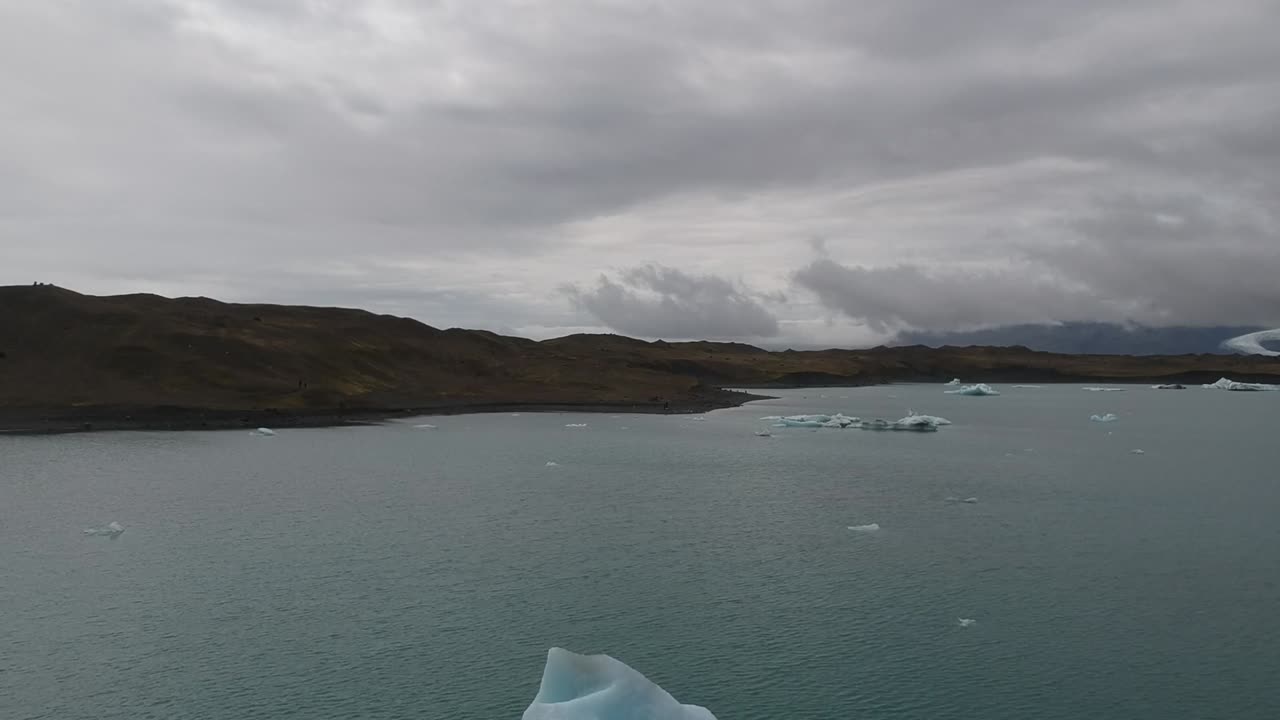 drone fly close to a giant glaciar iceberg floating on lagoon with mountains landscape at distance and cloudy dramatic sky.