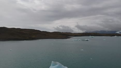 drone fly close to a giant glaciar iceberg floating on lagoon with mountains landscape at distance and cloudy dramatic sky.