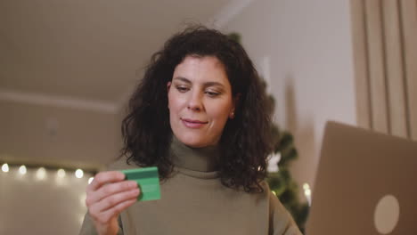 vista inferior de una mujer morena comprando en línea con una tarjeta de crédito usando una laptop sentada en una mesa en una habitación decorada con un árbol de navidad