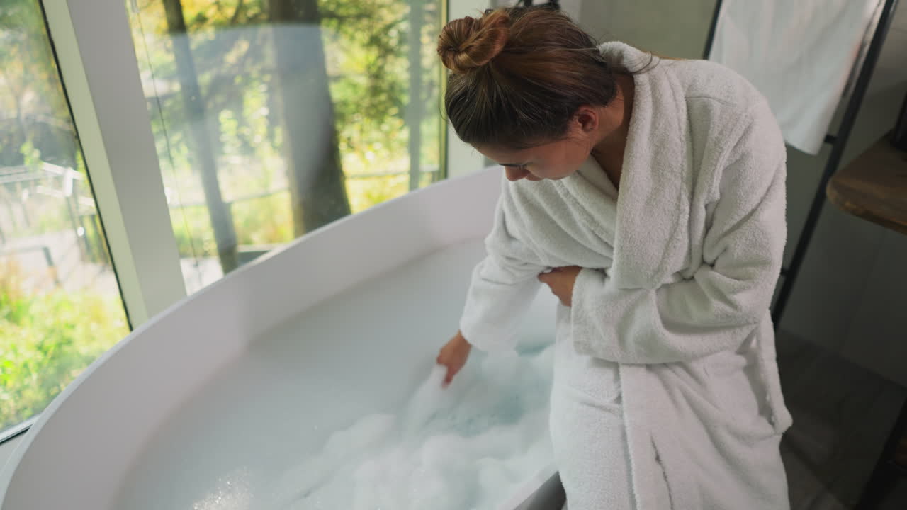 Woman playfully touches froth in bathtub. Lady in terry bathrobe stirs bubble foam checking temperature of water. Preparation for spa procedure