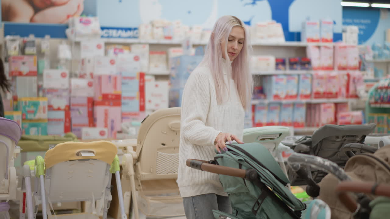Stroller pusher inspects baby stroller cover, opens and closes canopy while another female shopper passes by with shopping basket in bright retail aisle with blurred baby products