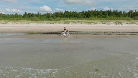 vista aérea con una joven de cabello largo de pie con una bicicleta en la playa de arena, día soleado, playa de arena blanca, concepto de estilo de vida activo, baja órbita ancha de drones