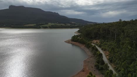 Sobrevuelo-Aéreo-Tranquilo-Lago-Cazador-Con-Reflejo-Del-Sol-Y-Carretera-Costera-En-El-Costado