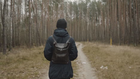 hombre caminando por un sendero forestal con una mochila, una chaqueta y un sombrero para el frío, persona sola e irreconocible vista desde atrás