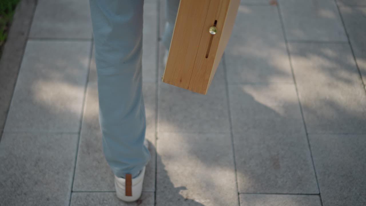 sneakers walking on tiled pavement, courier carrying wooden painting case under dappled shade rear view of jeansclad legs and confident stride, long soft shadows and textured tiles, purposeful
