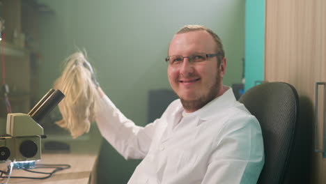 una vista de cerca de un técnico juguetón en una bata de laboratorio blanca y una peluca rubia, rascándose la cabeza con una mano de guante negro mientras sonríe a la cámara