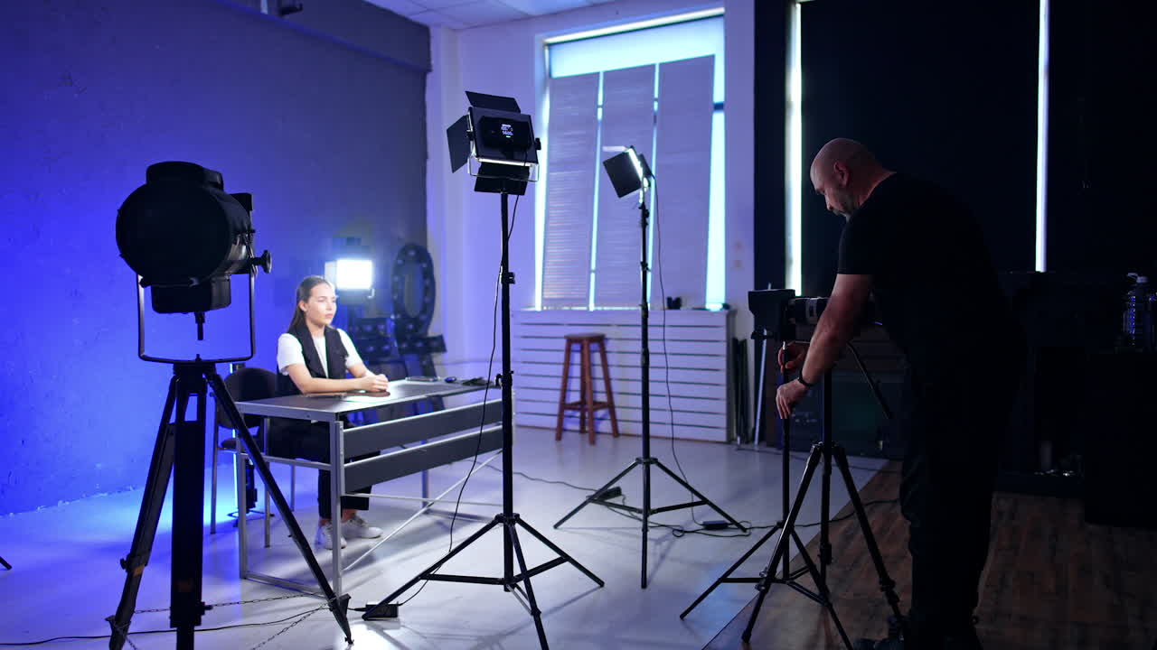 Interior of a modern photo studio where preparation for footage is taking place. Brunette girl sits at desk. Cameraman arranges necessary equipment.