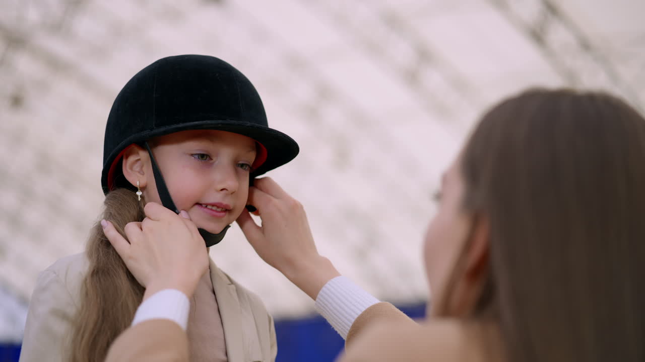 Brunette lady fits jockey's hat to a young girl's head. Woman ties the strap of a hat on child's head. Low angle view. Blurred backdrop.