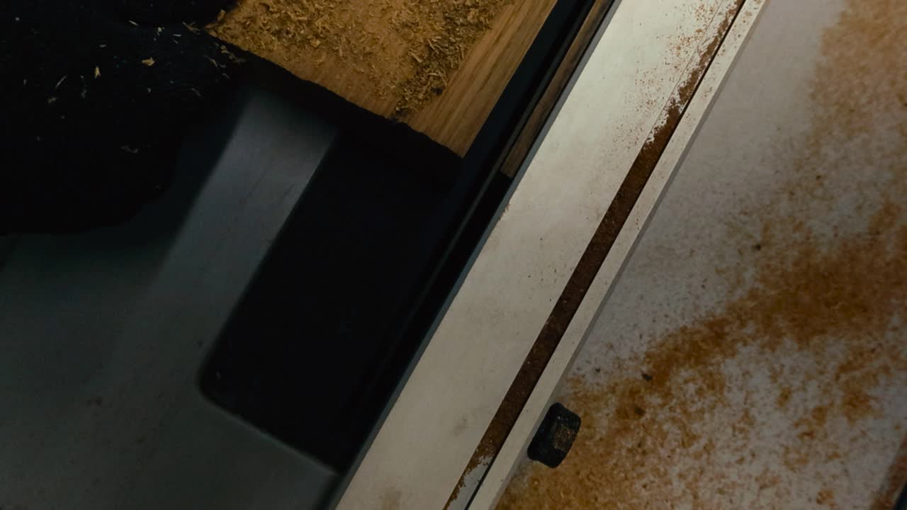 Top down slow motion video of a hand pushing and sawing a oak wooden board or slab in a carpentry workshop while sawdust and woodchips fly in slow motion. Gloved black hand pushing the wood forward