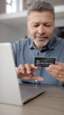 vertical video of senior biracial man using laptop and using credit card at home, vertical