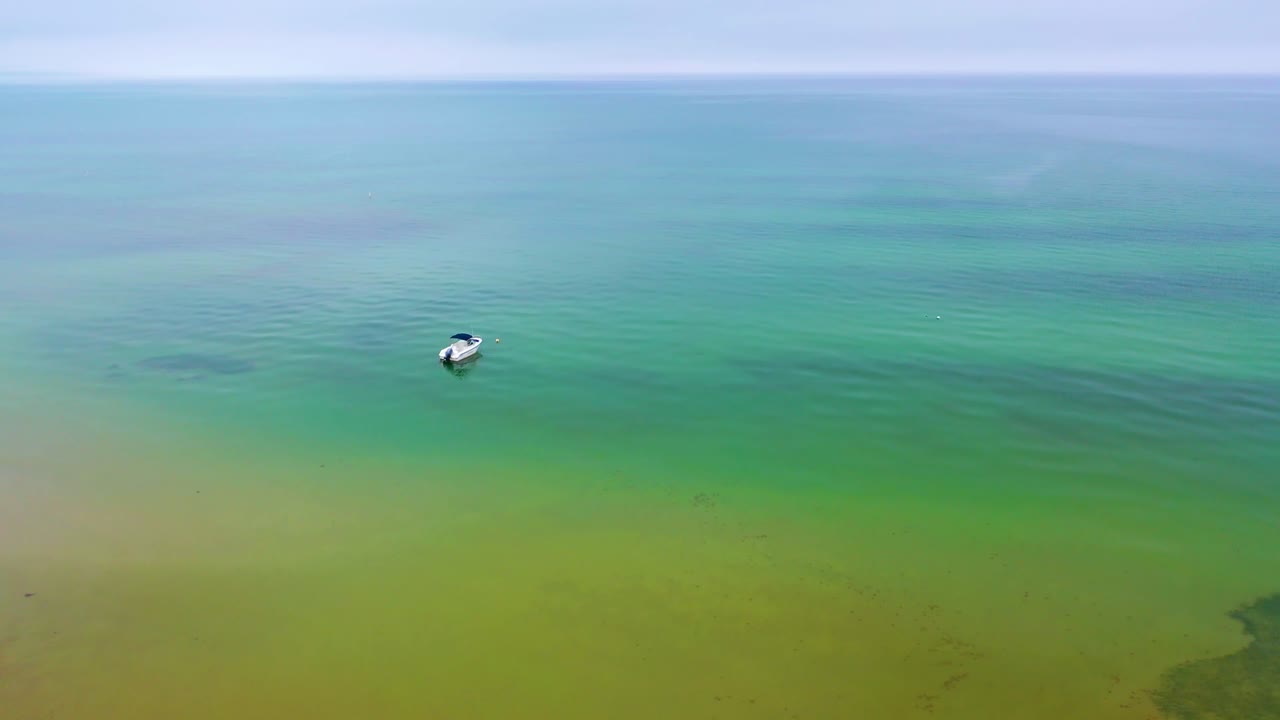 Drone video overlooking a single boat anchored in tranquil green waters, surrounded by gentle ripples and framed by a wide, misty horizon beneath an overcast sky