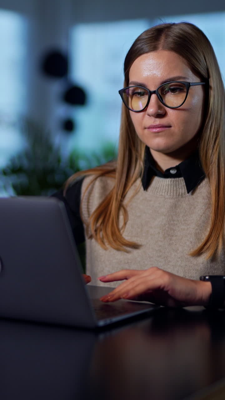 Blonde woman in glasses opens laptop at the table in front of her. Beautiful young woman working at computer in the office. Blurred background. Vertical video