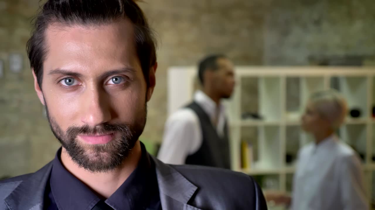 apuesto hombre de negocios confiado con barba está viendo en la cámara en la oficina, los colegas están hablando en el fondo, concepto de comunicación