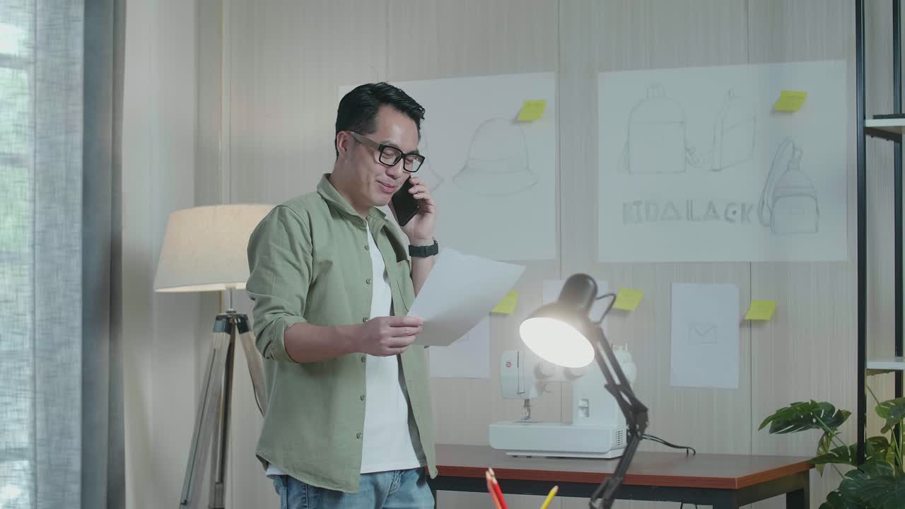 Desainer Pria Asia Sedang Menelepon Menggunakan Ponsel Pintar Sambil Melihat Desain Tata Letak di Tangan dan Berjalan di Kantor