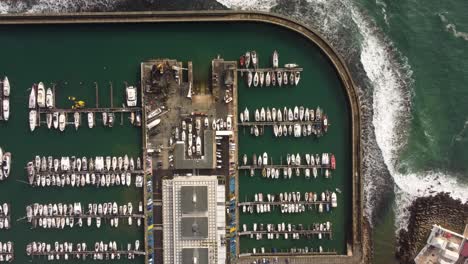 Aerial-top-down-minimalist-view-of-yacht-sail-boat-moored-at-harbor-with-ocean-waves-crashing-on-stone-pier-port