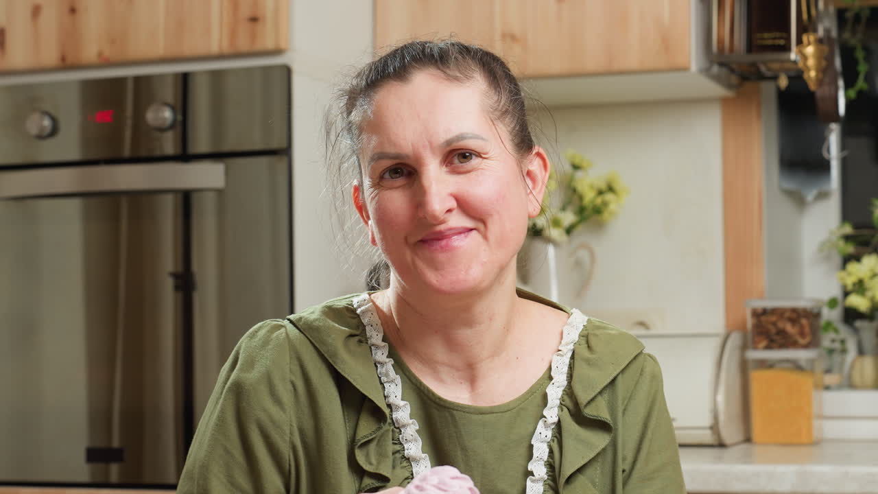 Portrait view of cheerful woman in green dress smiling warmly while holding cupcake in home kitchen with soft lighting, surrounded by flowers, wooden cabinets, and cozy kitchen decor elements