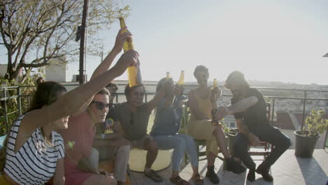 happy friends raising bottles of beer up and toasting while sitting on rooftop during an outdoor party