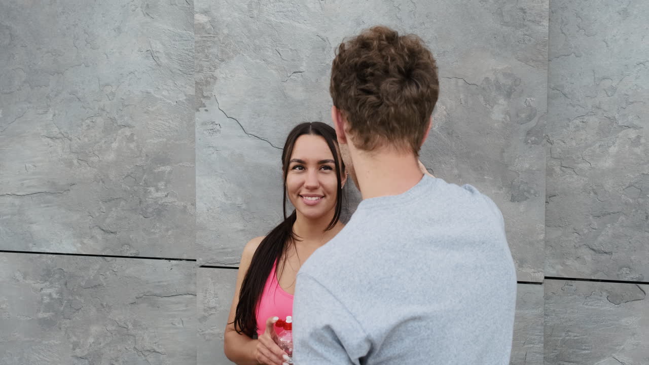 linda corredora apoyada contra una pared, bebiendo agua y hablando con su novio parado frente a ella mientras se toman un descanso durante la sesión de entrenamiento en la ciudad