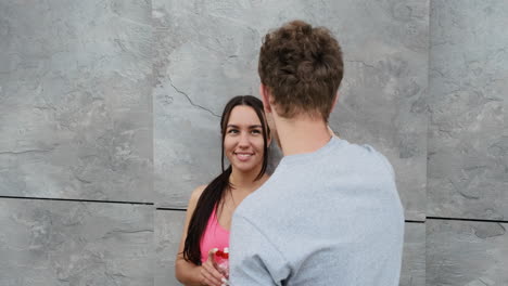 Pretty-Runner-Girl-Leaning-Against-A-Wall,-Drinking-Water-And-Talking-To-Her-Boyfriend-Standing-In-Front-Of-Her-While-They-Are-Taking-A-Break-During-Training-Session-In-The-City