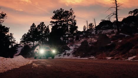 stunning tracking shot of a blue suv with bright headlights driving along a snowy dirt mountain road during sunset