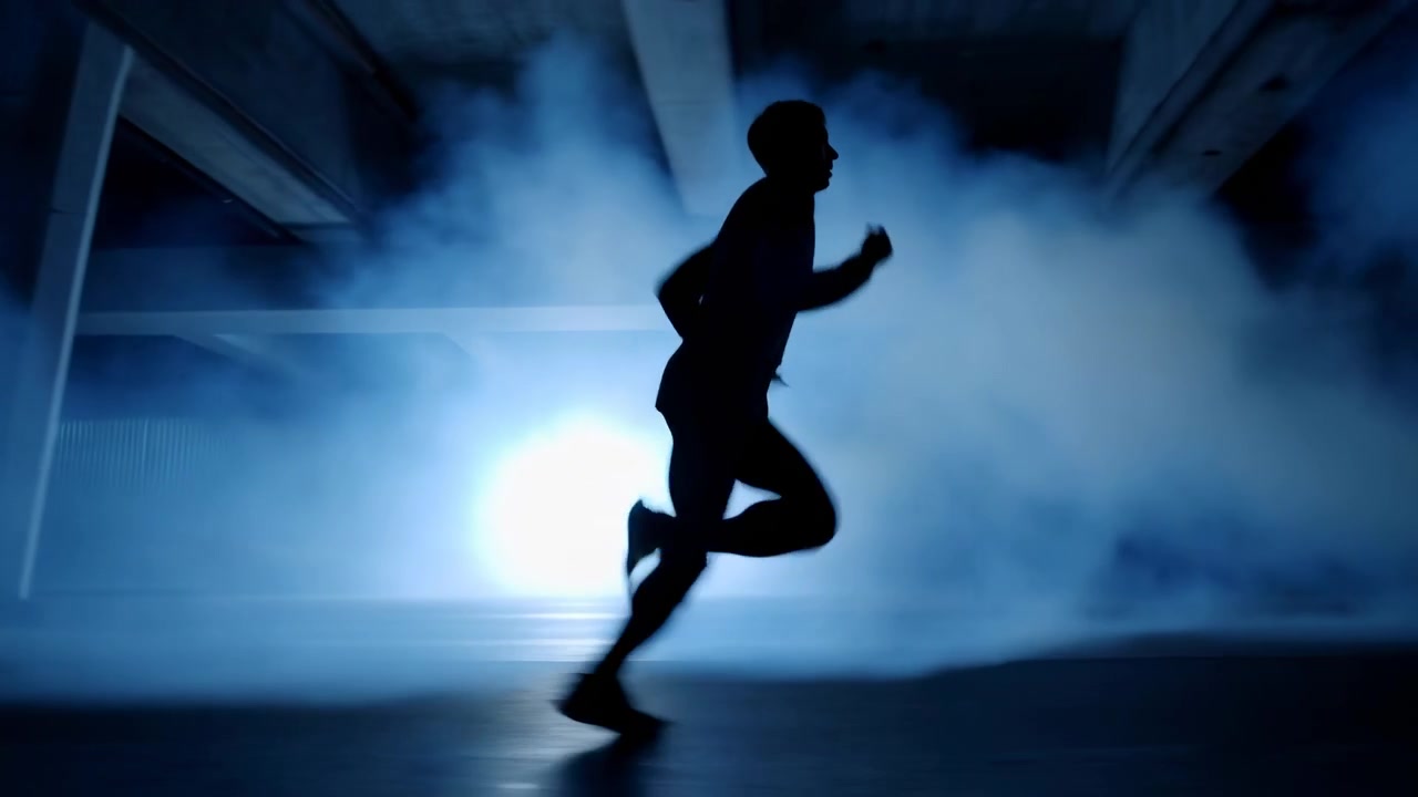 Lone silhouetted runner moves through spacious industrial interior with blue haze, bright backlight, concrete columns, and shadowed floor, creating moody urban scene with strong contrast and active
